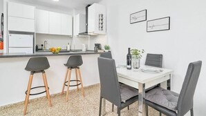 Et moderne køkken med lyse skabe, en sort bordplade og et spiseområde med et hvidt bord og grå stole.
