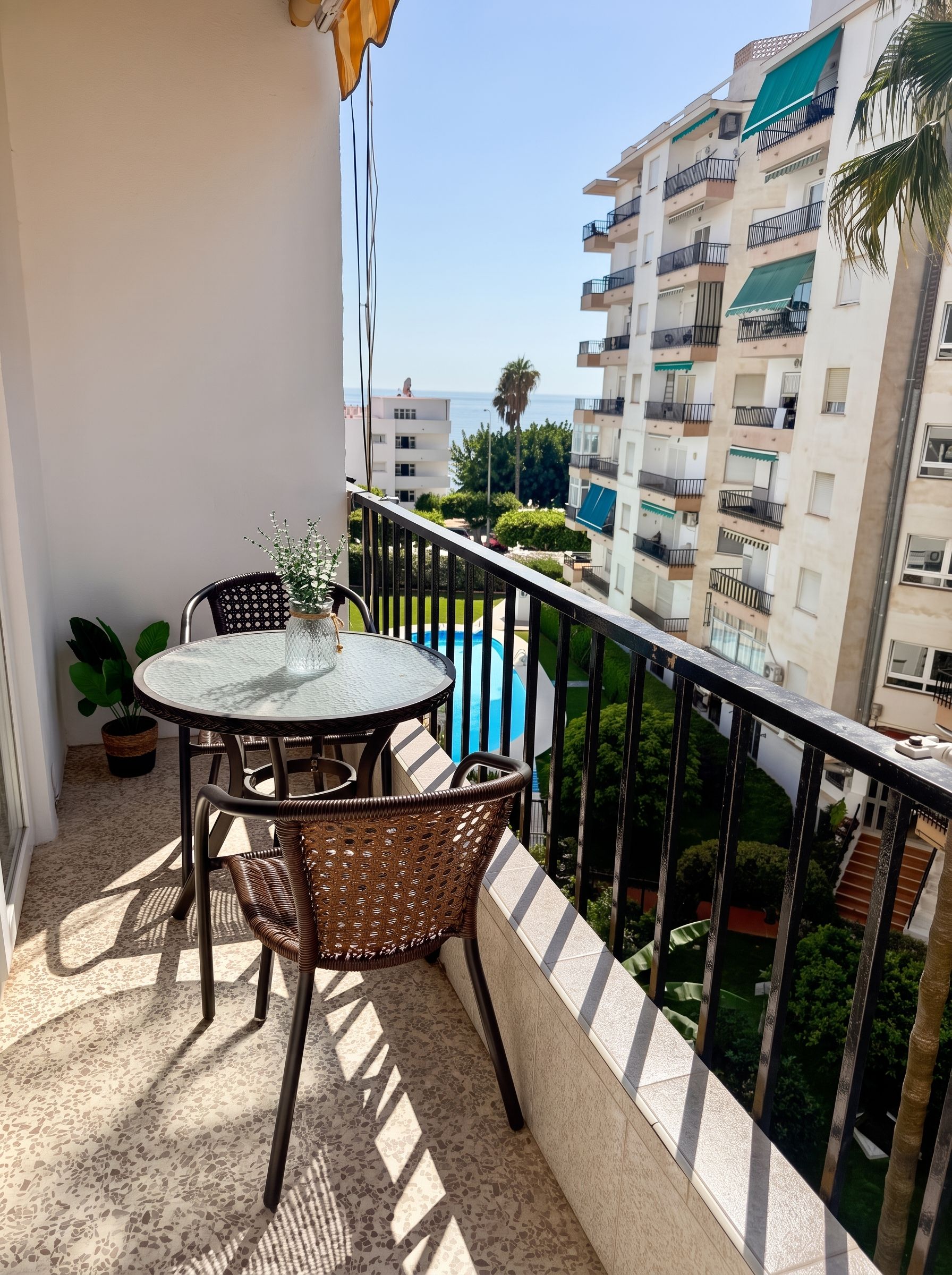 Un balcon doté d’une table ronde et de chaises en osier, offrant une vue sur une piscine et les bâtiments adjacents.
