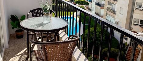 Un balcon doté d’une table ronde et de chaises en osier, offrant une vue sur une piscine et les bâtiments adjacents.