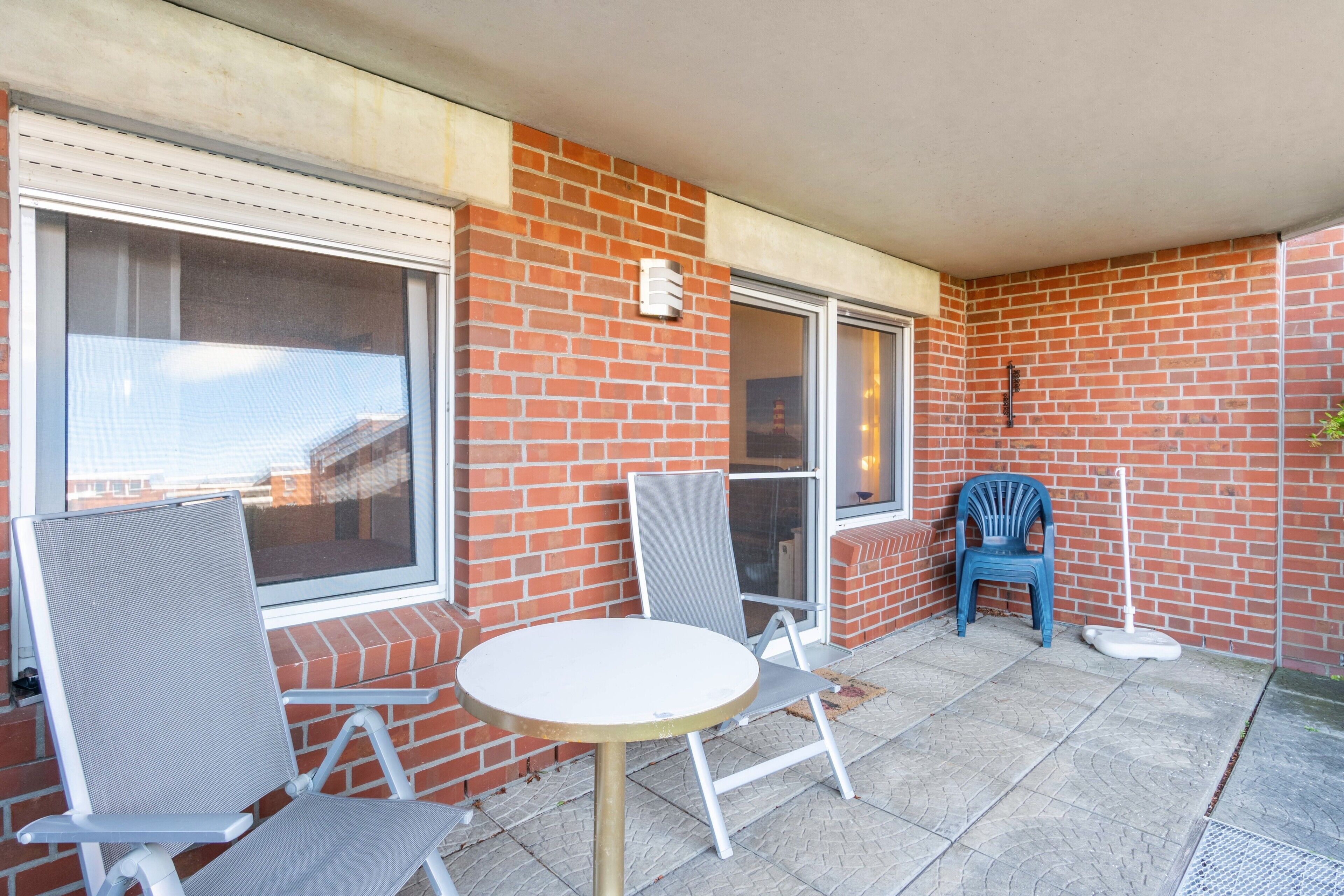 Un patio con una mesa redonda y dos sillas, una silla de plástico azul y una escoba apoyada contra la pared.