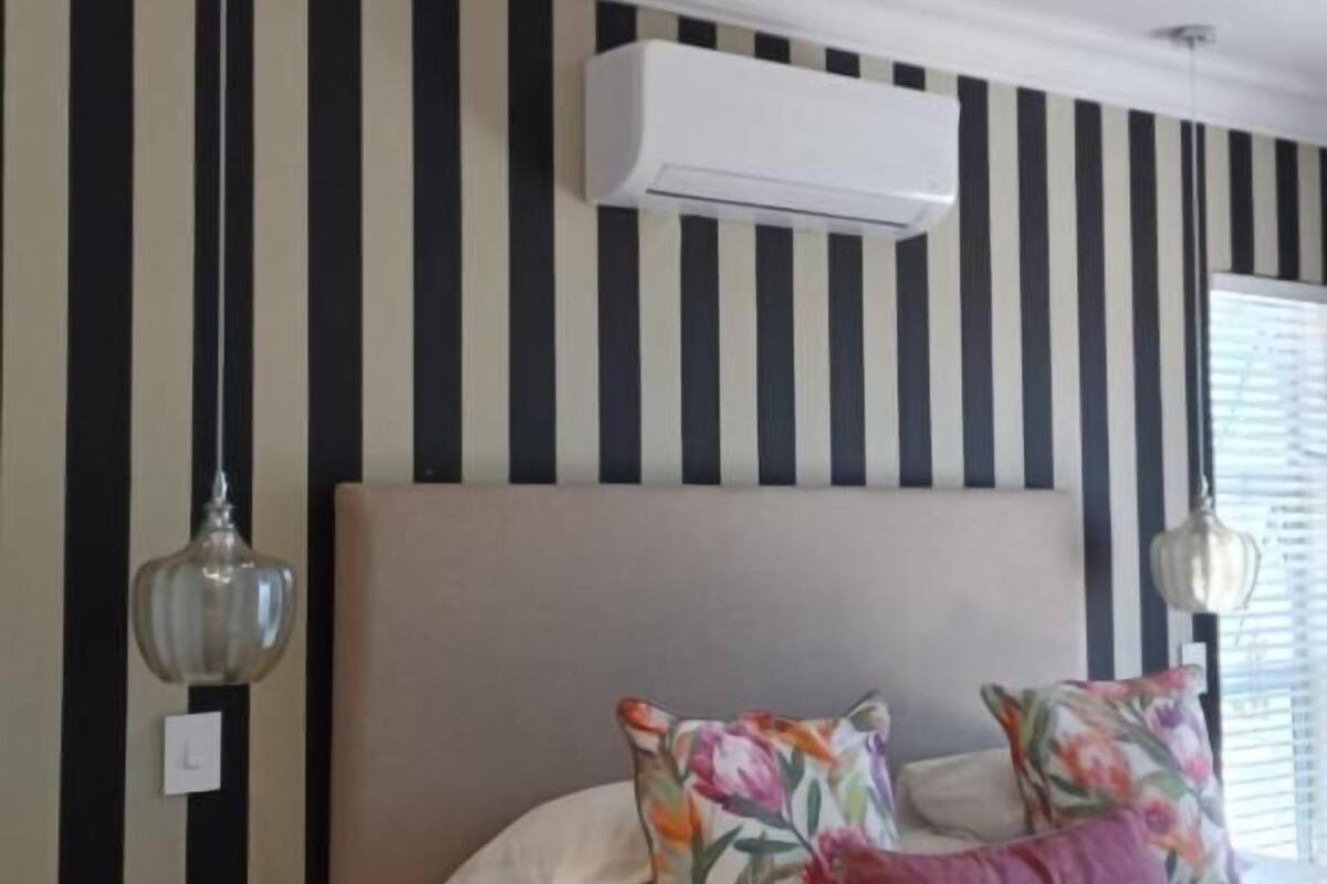 Un dormitorio con pared a rayas, una cama con almohadas estampadas y lisas, una mesita de noche con una lámpara y un equipo de aire acondicionado.