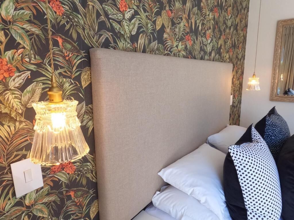 A bedroom with a floral wallpaper, a headboard, a bedside lamp, and a mirror.
