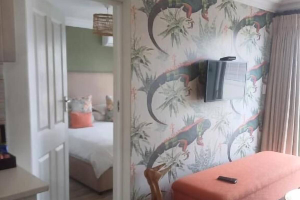 Una habitación con cama, una mesita, un banco, un televisor montado en la pared y una puerta que da a otra estancia.