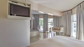 Ein Hotelzimmer mit Bett, Wand-Fernseher, einer Sitzecke mit Sofa und Sesseln sowie großen Fenstern mit Vorhängen.