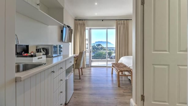 Cocina con armarios blancos, microondas y pileta. Hay una silla y una cama en la habitación, con vista al exterior a través de un ventanal grande.