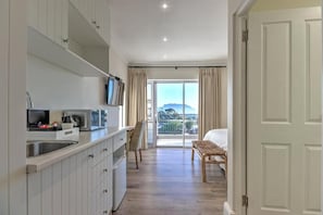 A kitchen with white cabinets, a microwave, and a sink. There is a chair and a bed in the room, with a view of the outdoors through a large window. - Thyme Wellness Spa and Guesthouse (Cape Town)