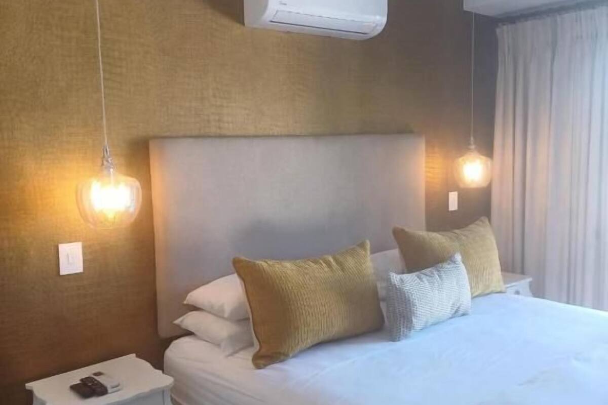 Habitación de hotel con cama, mesitas de noche, unidad de aire acondicionado e iluminación en la pared.