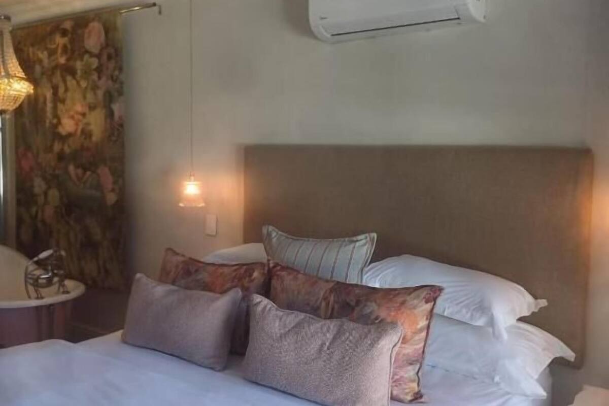 Una cama bien hecha con varios almohadones, un aire acondicionado de pared y una mesita de noche pequeña.