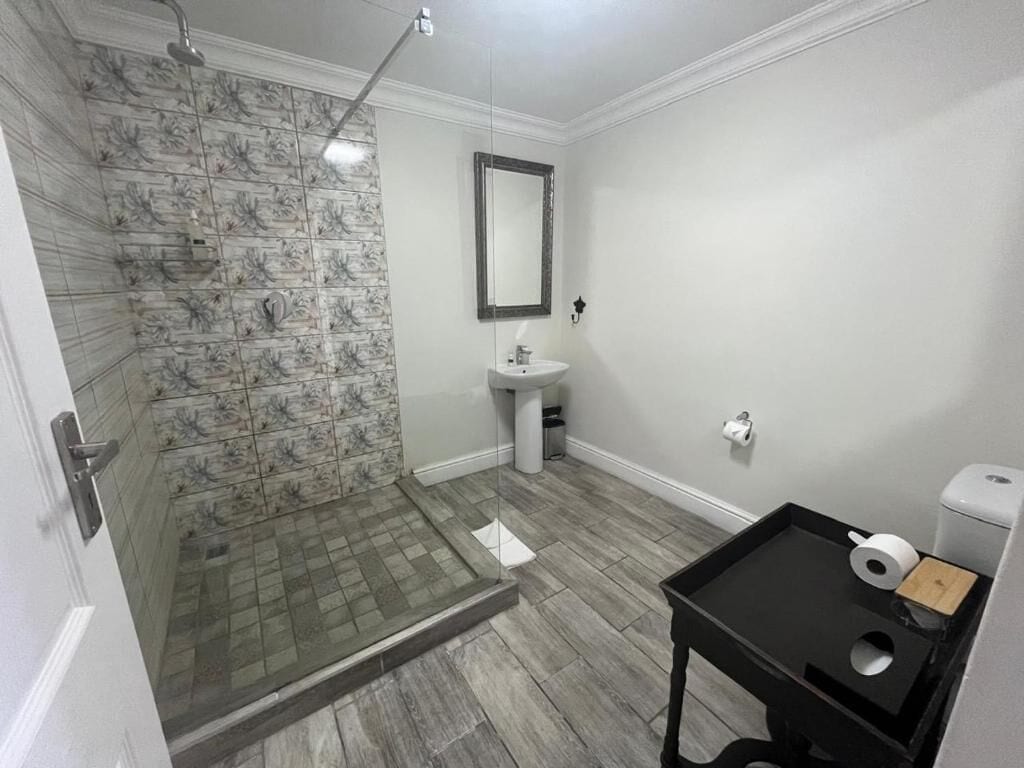 A bathroom with a shower, sink, mirror, and a small table with a toilet paper holder.
