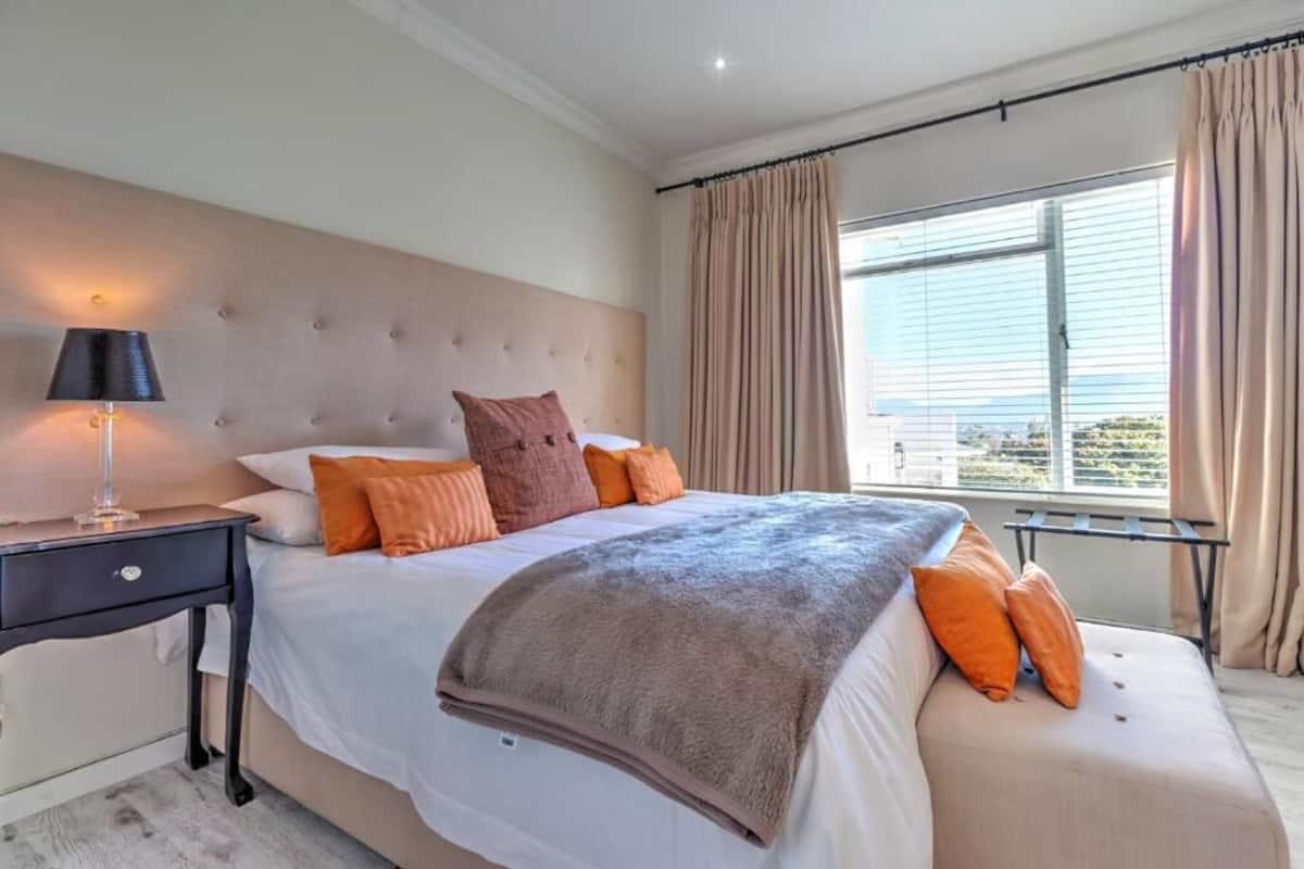 Un dormitorio con una cama grande, una mesita de noche con lámpara, una ventana con cortinas y vistas al exterior.