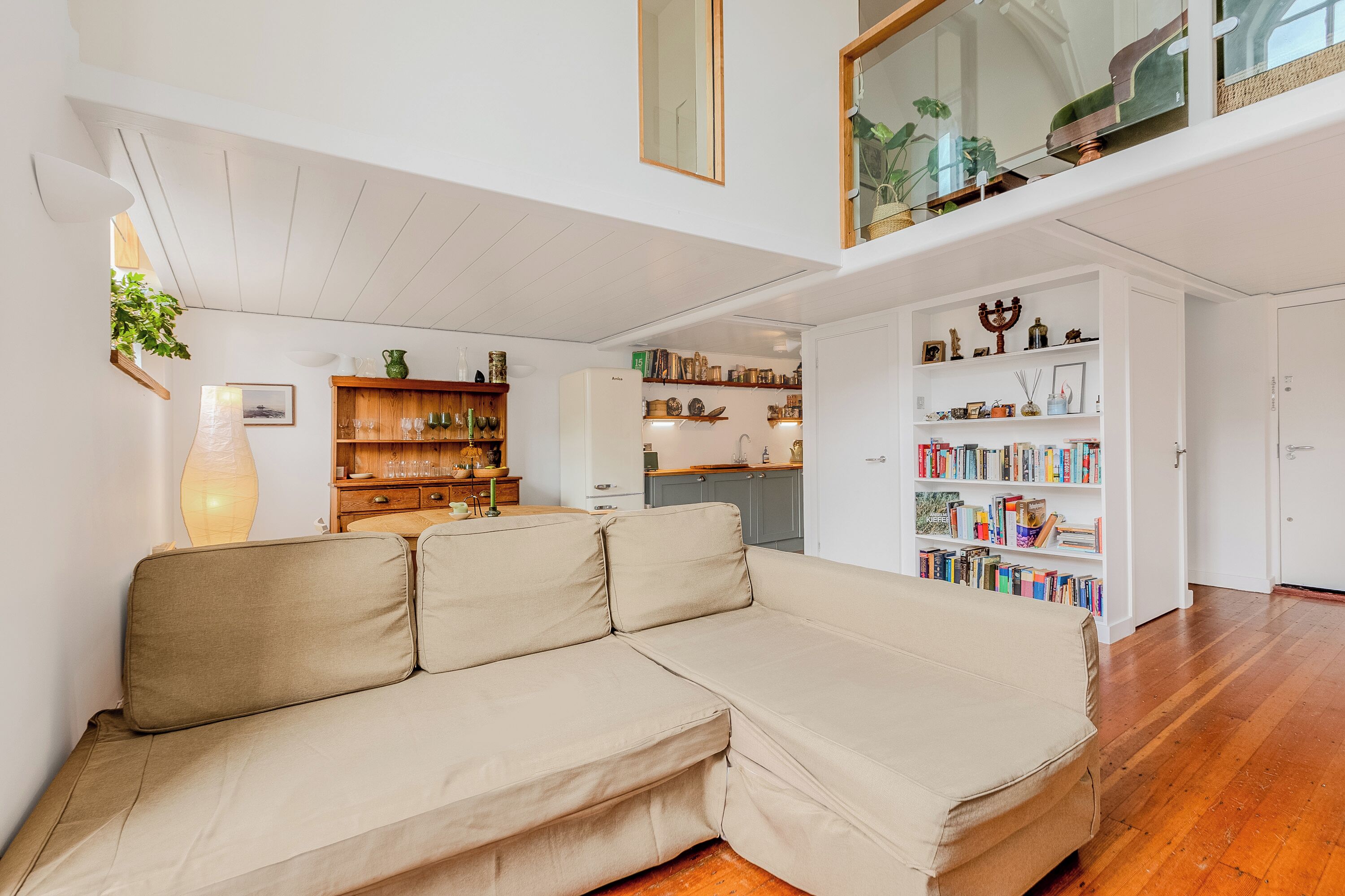 En moderne stue med en beige hjørnesofa, træhylder med bøger og dekorative genstande, og et køkkenområde i baggrunden.