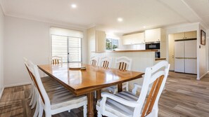 A spacious dining area with a wooden table and white chairs, a kitchen with white cabinets and appliances, and a view of the outdoors through the windows.