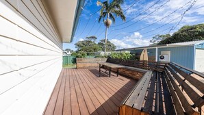A wooden deck with benches, a palm tree, and a building with a blue and white exterior. - Cookies Hideway - Coastal holiday vibes. (WOOLI)