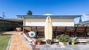 Une maison avec un patio couvert, une planche de surf appuyée contre la clôture et des plantes en pot.