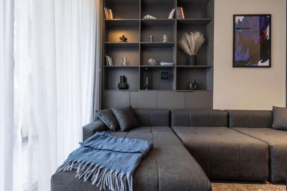Uma sala de estar moderna com um sofá modular cinza, uma estante com diversos objetos decorativos e uma pintura abstrata emoldurada na parede.