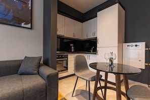 A compact living space with a sofa, a dining table with two chairs, a kitchen area with cabinets and appliances, and a refrigerator. - Prishtina City Apartments 2 (Pristina)