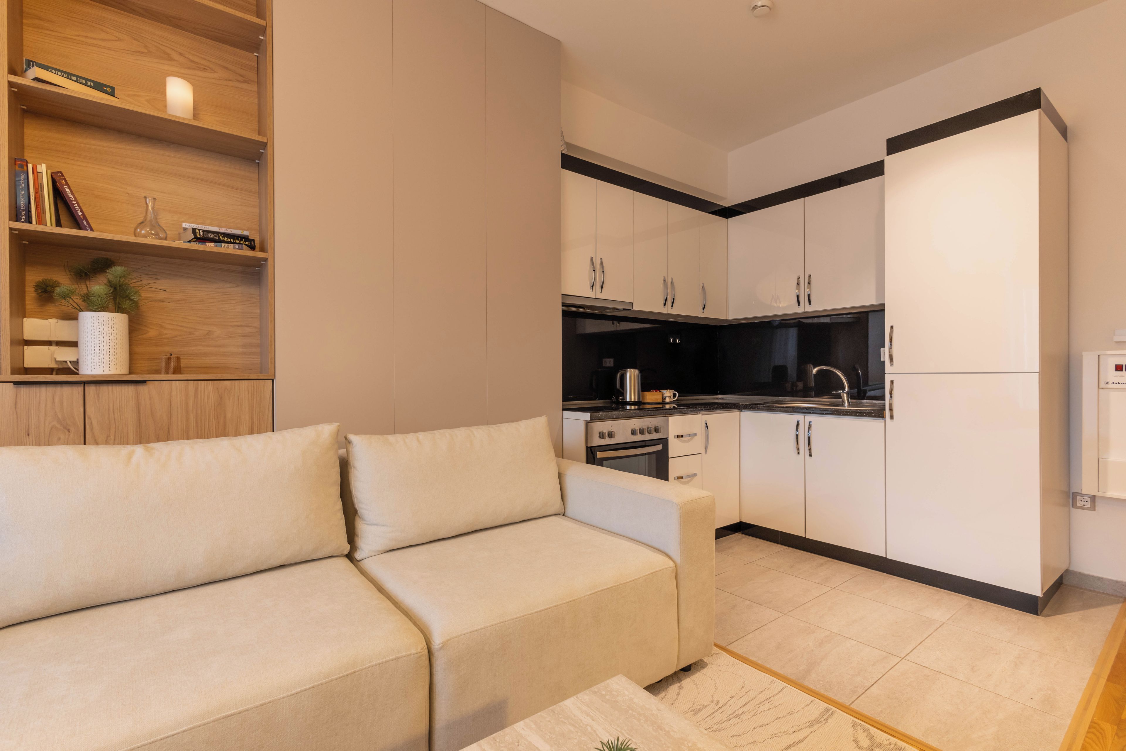 A modern living room with a beige sofa, a coffee table, and a kitchen area with white cabinets and a black countertop.