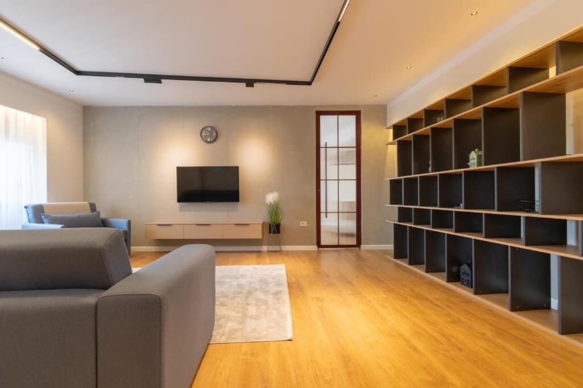 Uma sala de estar moderna com um sofá cinzento, uma televisão de tela plana, piso de madeira e estantes embutidas.