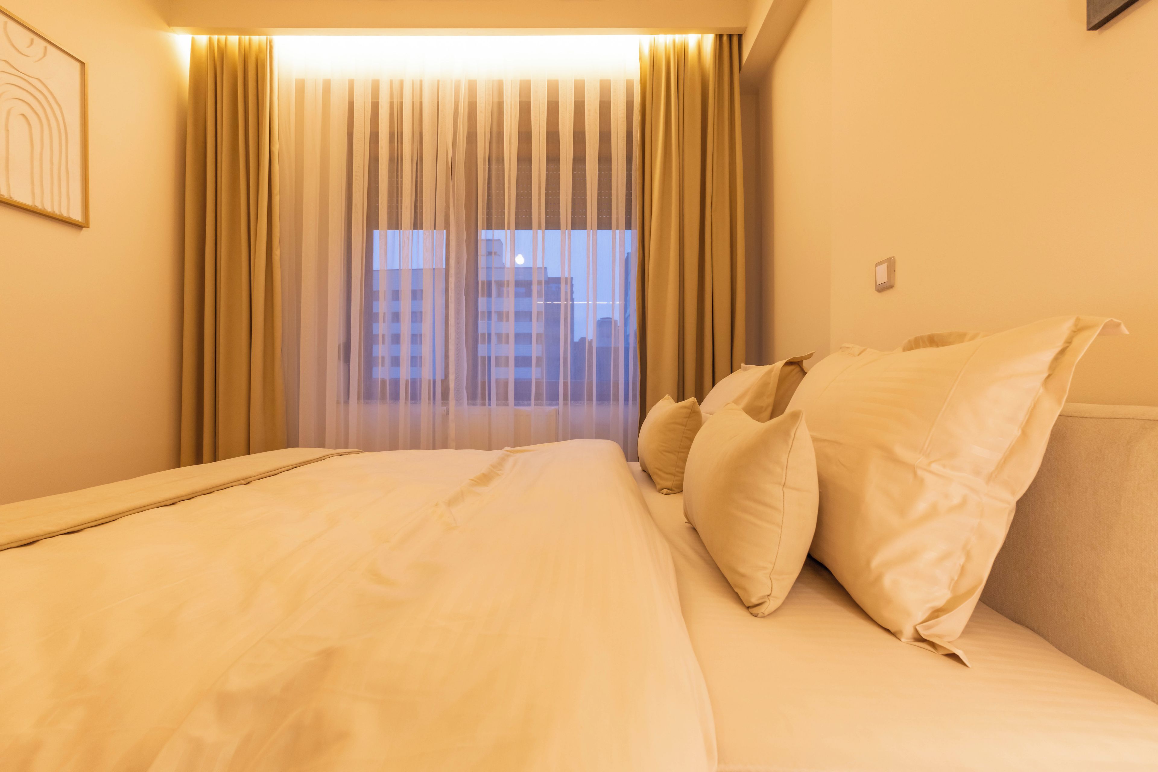 Habitación de hotel con una cama grande, cortinas color beige y vista a edificios por la ventana.