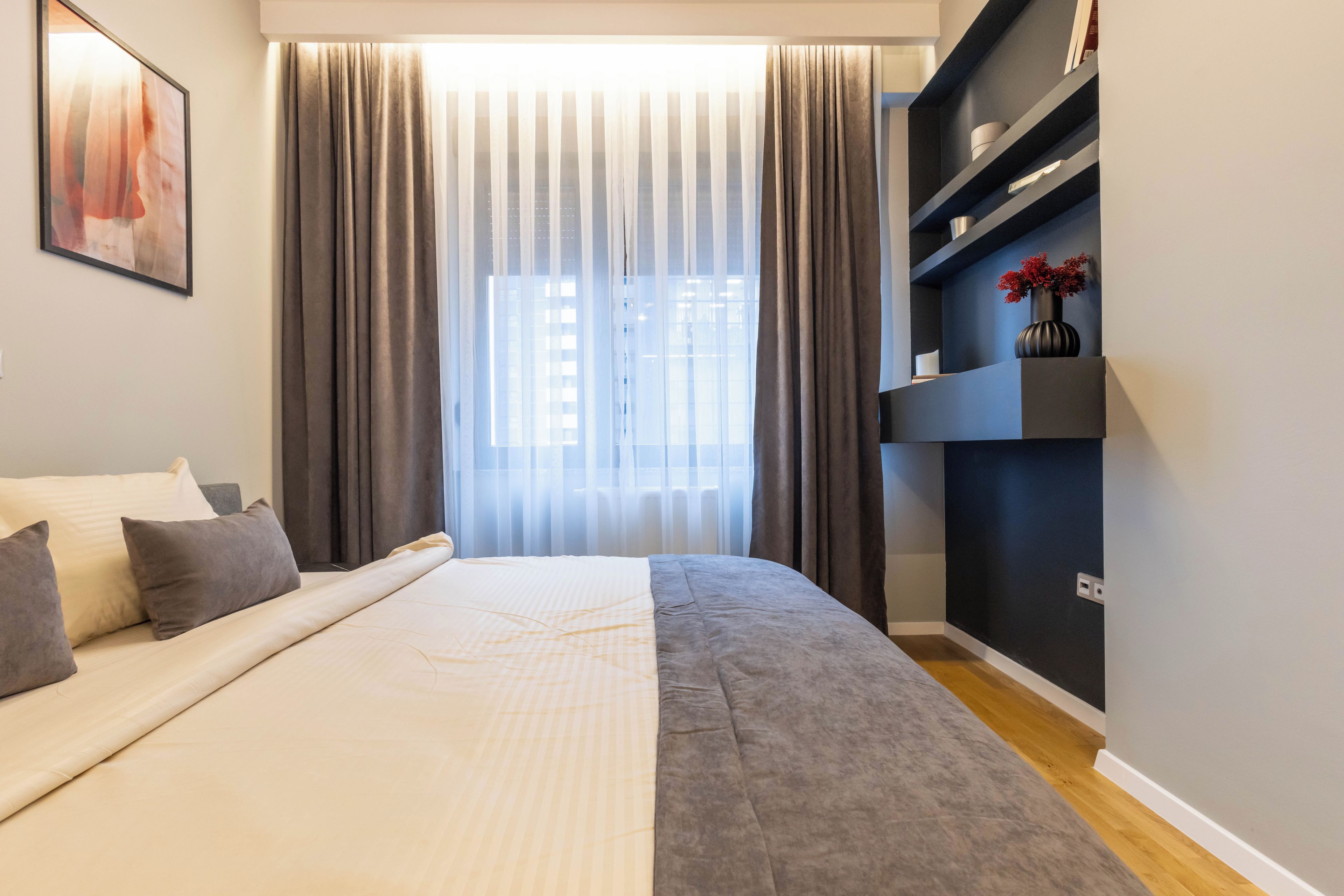 A modern bedroom with a large bed, a bedside table, a built-in shelf with decorative items, and a window with curtains.