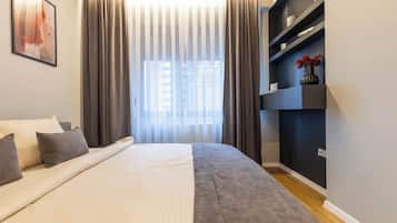 A modern bedroom with a large bed, a bedside table, a built-in shelf with decorative items, and a window with curtains.