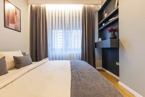 A modern bedroom with a large bed, a bedside table, a built-in shelf with decorative items, and a window with curtains. - Prishtina City Apartments 2 (Pristina)