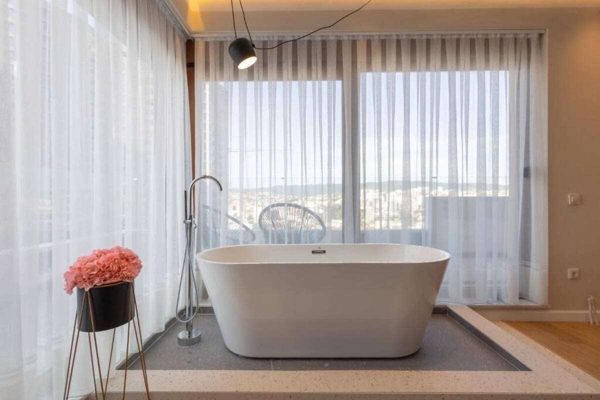Un baño moderno con una bañera independiente, un box de ducha con paredes de cristal y vistas a un paisaje urbano a través de amplios ventanales.
