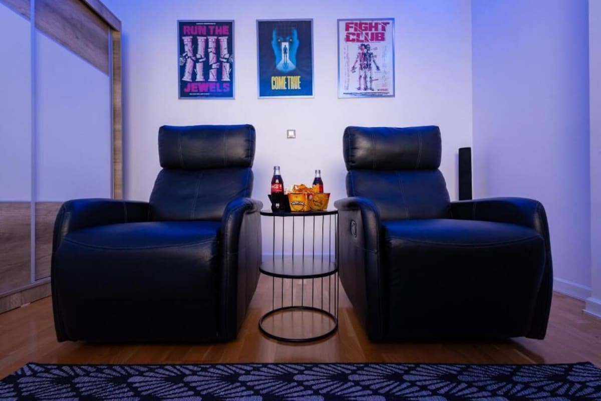 Um quarto com duas poltronas de couro preto, uma mesa redonda pequena com garrafas e petiscos, e três quadros nas paredes.