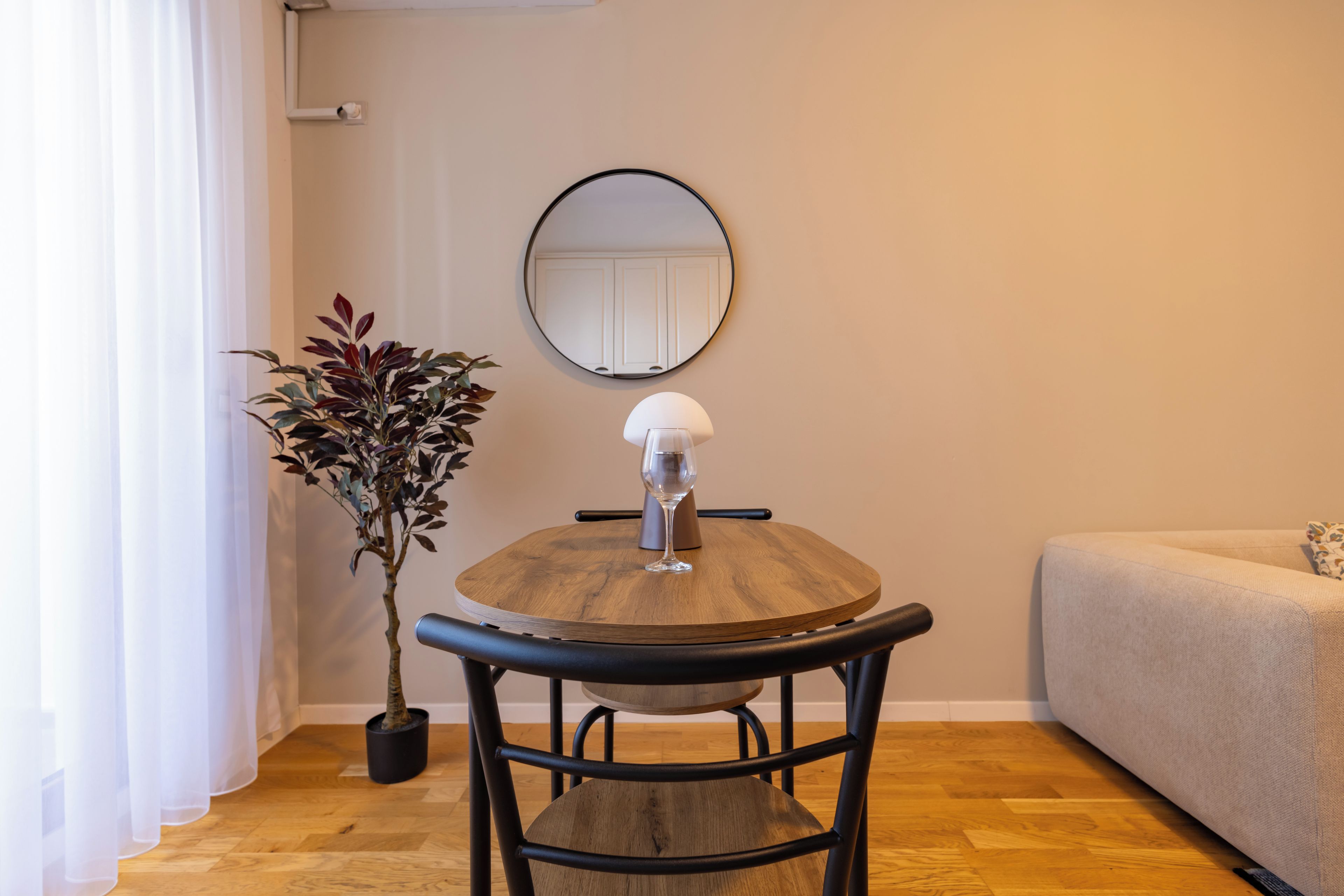 Una mesa redonda de madera con un vaso y una silla metálica, una planta en maceta, un espejo redondo en la pared y un sofá.