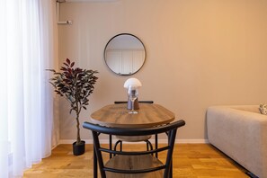 A round wooden table with a glass and a metal chair, a potted plant, a round mirror on the wall, and a sofa. - Prishtina City Apartments 2 (Pristina)