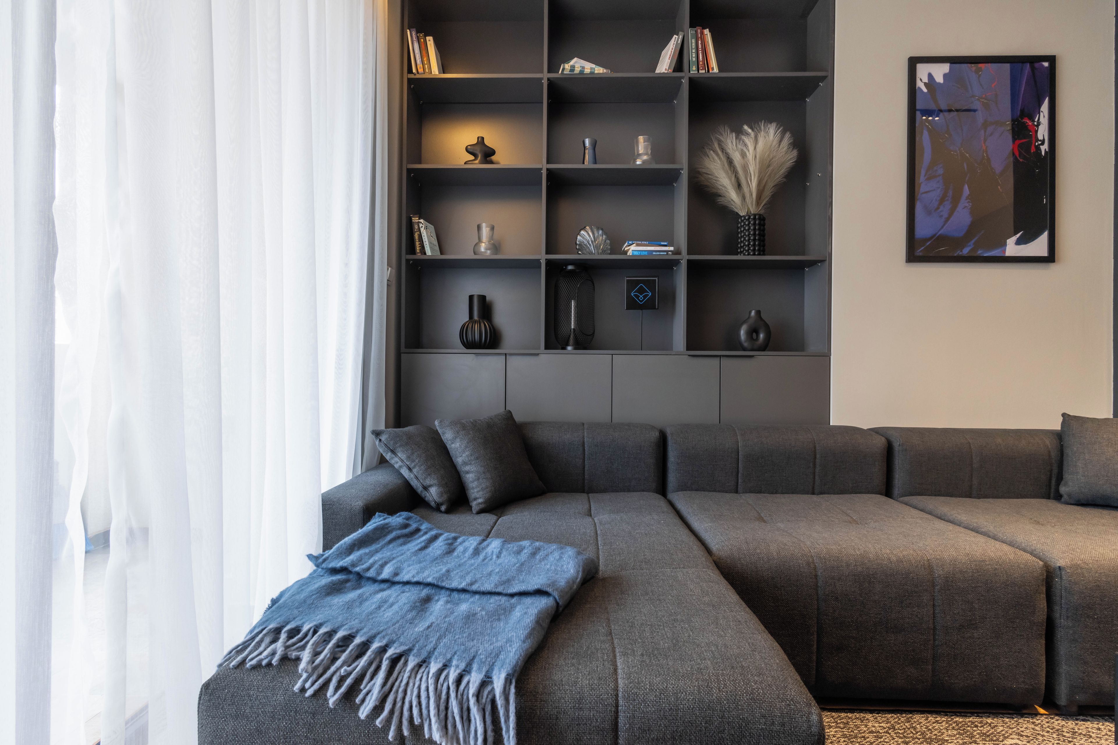 Un salon moderne avec un canapé d’angle gris, un plaid bleu et une bibliothèque agrémentée de divers objets décoratifs.