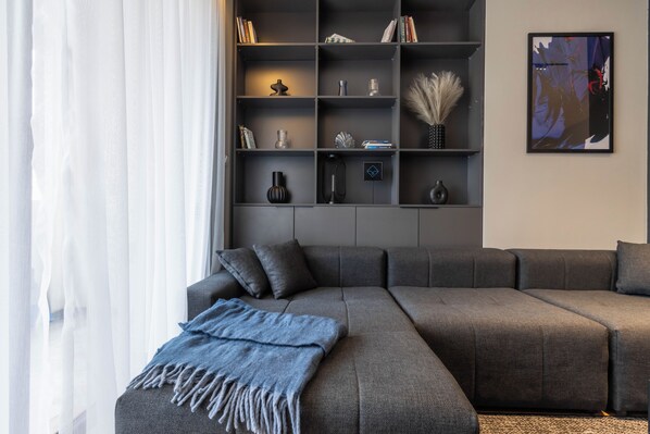 A modern living room with a grey sectional sofa, a blue throw blanket, and a bookshelf with various decorative items. - Prishtina City Apartments 2 (Pristina)