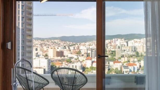 A modern bathroom with a freestanding bathtub, two wire chairs, and a city view through large windows. - Prishtina City Apartments 2 (Pristina)