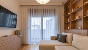 A modern living room with a sofa, a TV mounted on the wall, a bookshelf, and a large window with sheer curtains. - Prishtina City Apartments 2 (Pristina)