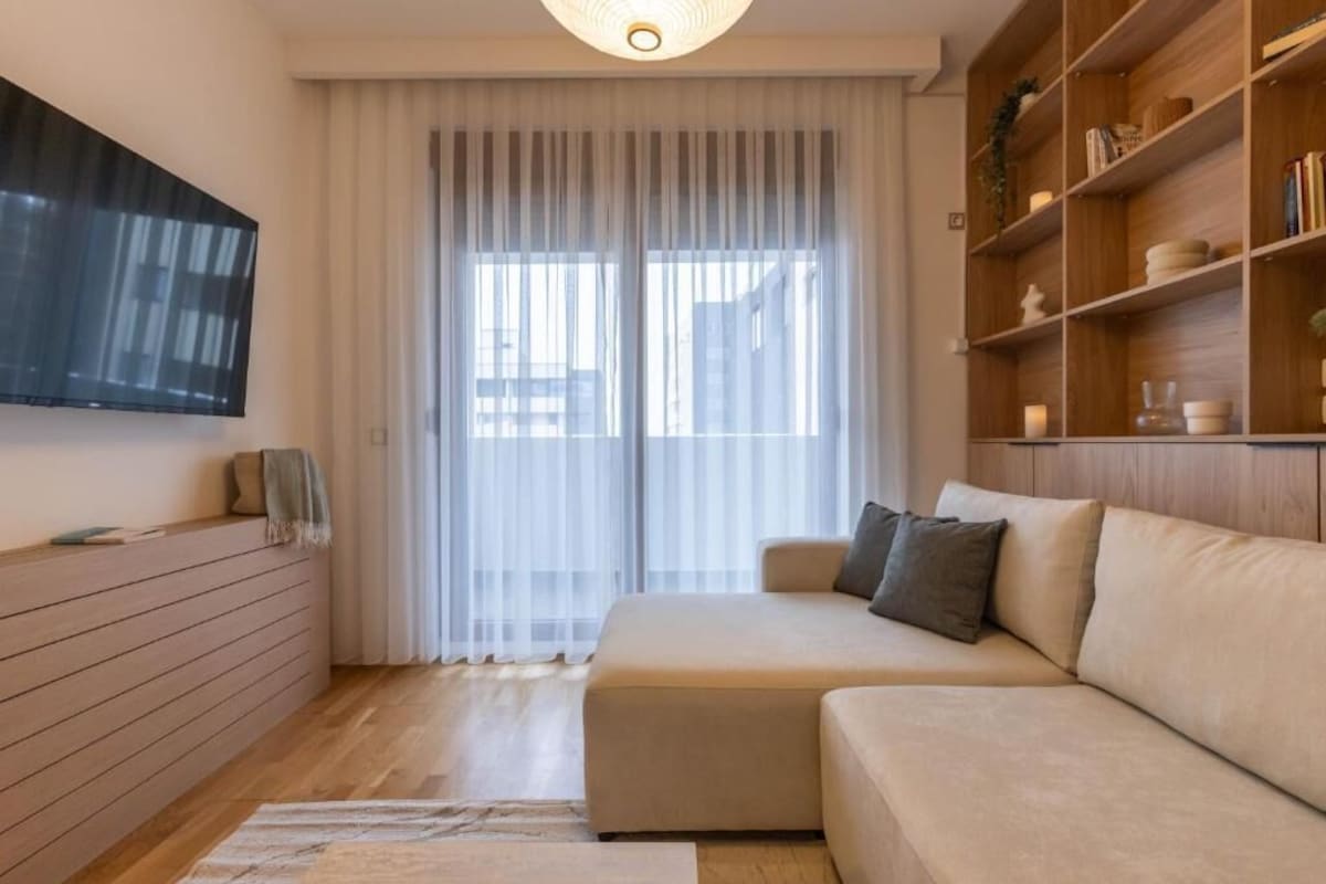 Uma sala de estar moderna com um sofá, uma televisão na parede, uma estante e uma janela grande com cortinas leves.
