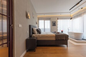 A modern bedroom with a large bed, bedside tables, a built-in bathtub, and a view of the city. - Prishtina City Apartments 2 (Pristina)