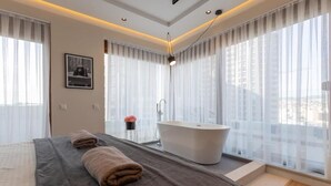 A modern bedroom with a large bed, a bathtub, and a city view through sheer curtains. - Prishtina City Apartments 2 (Pristina)