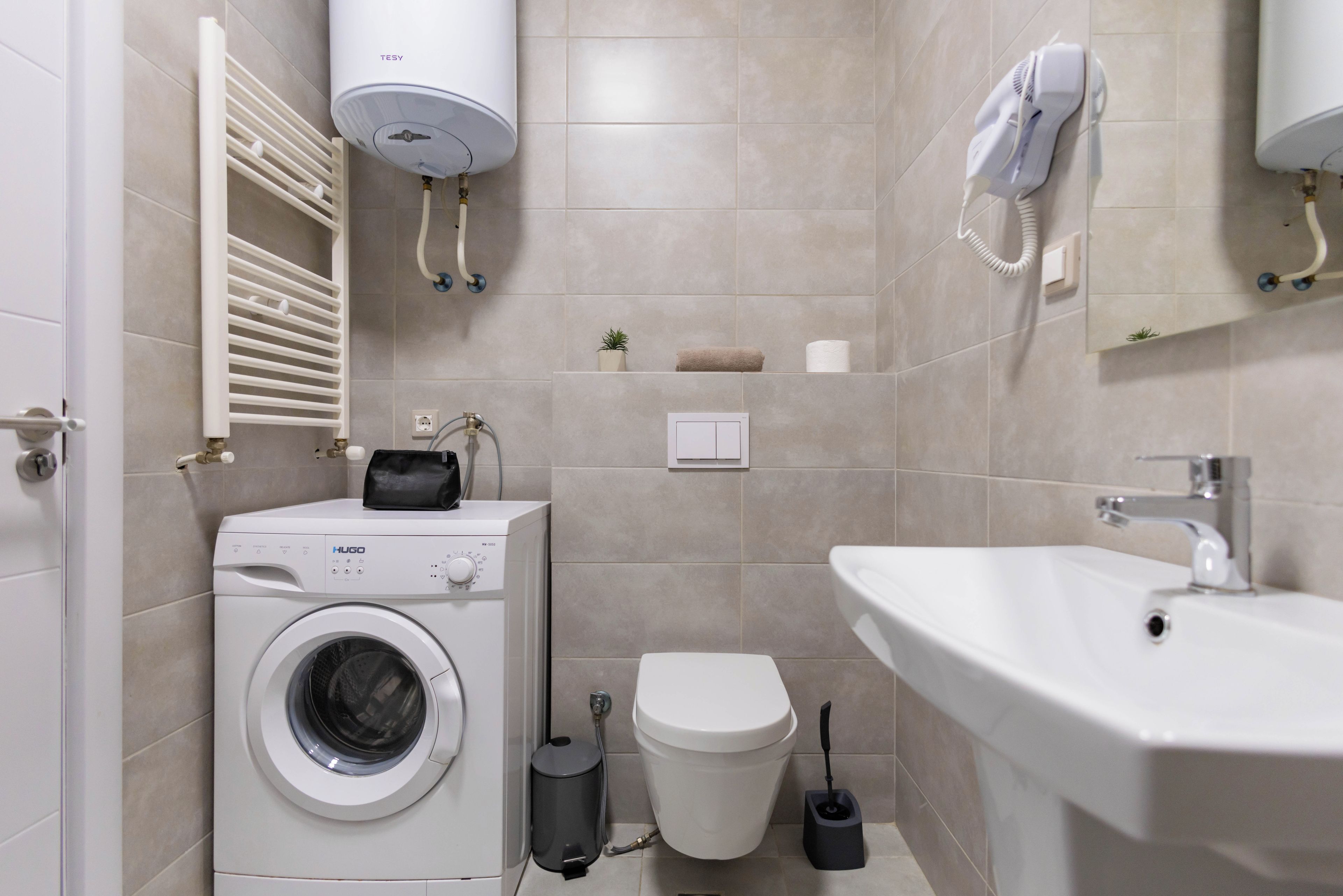 A compact bathroom with a washing machine, toilet, sink, towel rack, and a hairdryer.