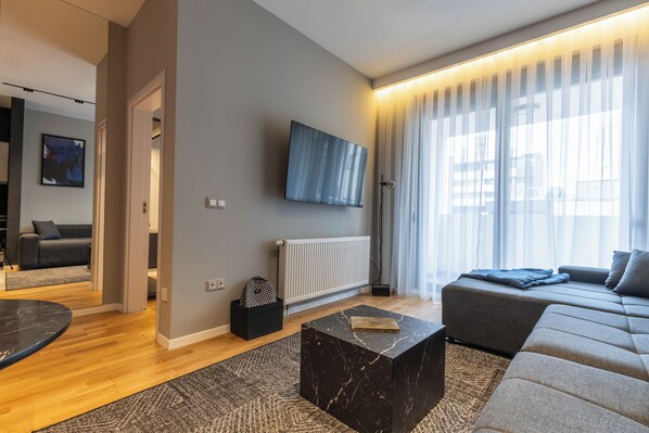 A modern living room with a grey sofa, a black coffee table, a mounted TV, and a large window with sheer curtains. - Prishtina City Apartments 2 (Pristina)