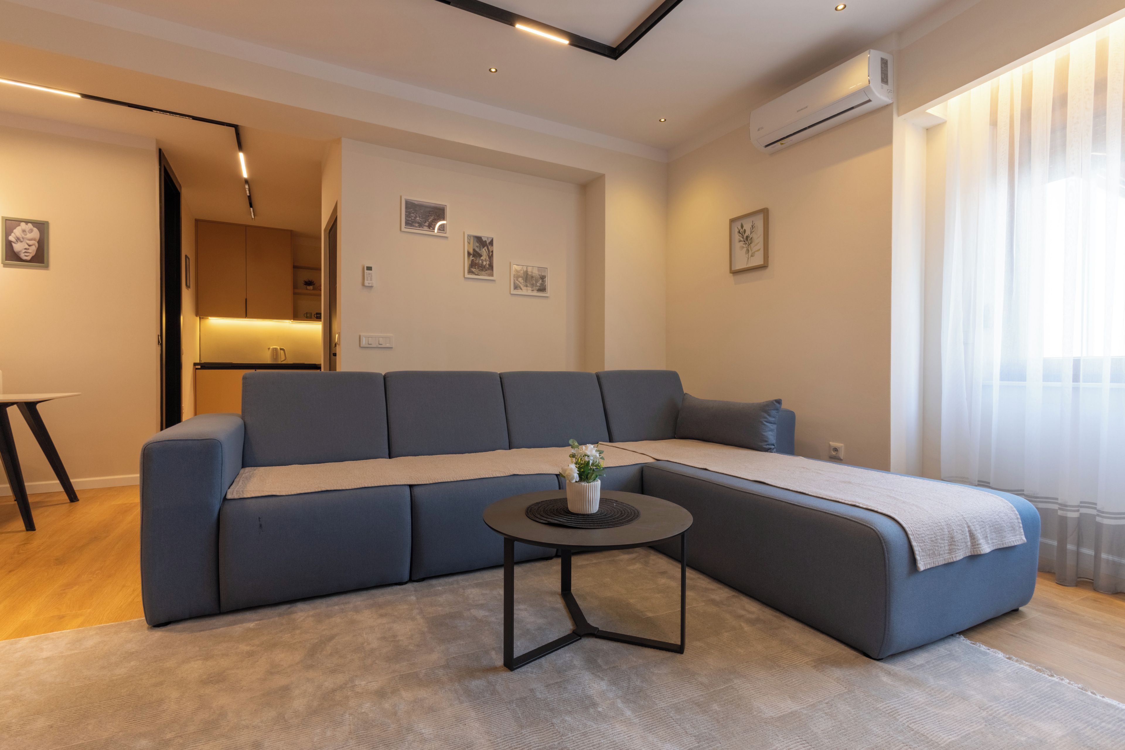 Una sala de estar moderna con un sofá seccional, una mesita redonda y una zona de cocina al fondo.