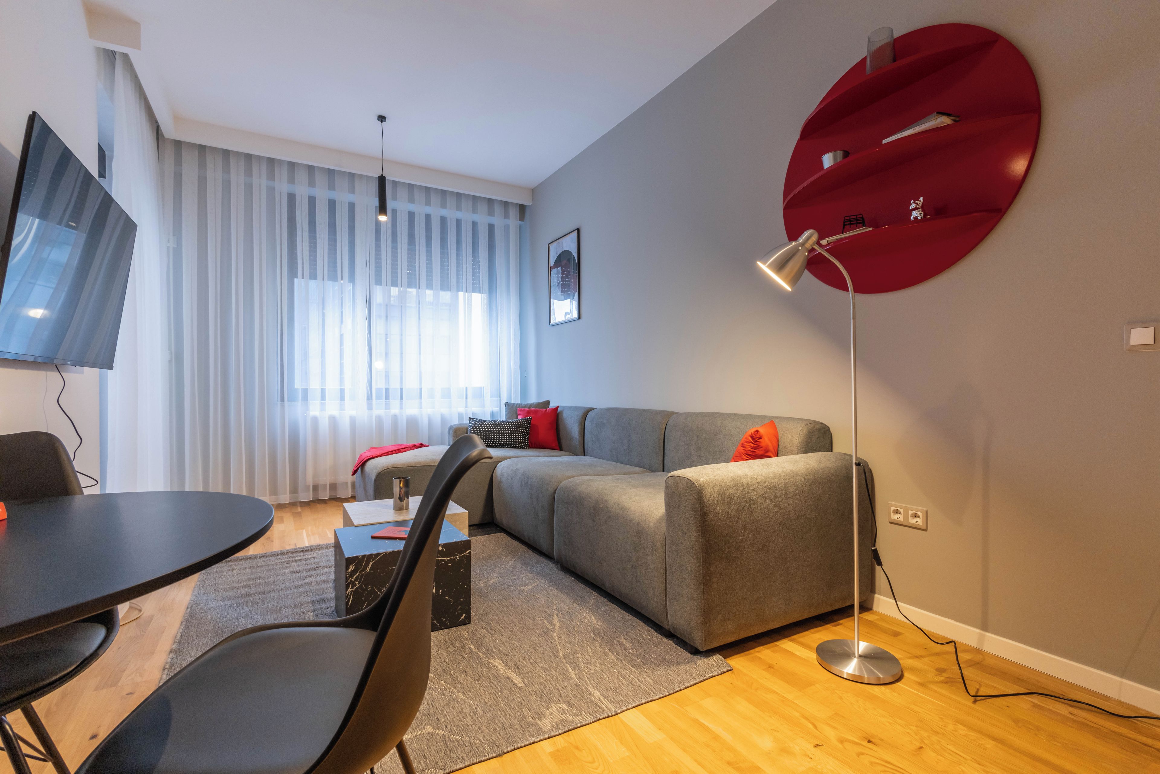 Een moderne woonkamer met een grijze bank, een ronde rode bijzettafel, een zwarte eettafel en een vloerlamp.