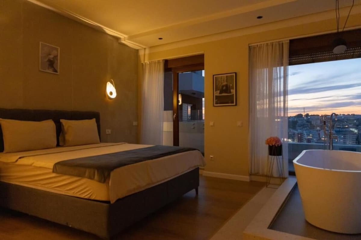 Quarto com cama, banheira, vista para a cidade e porta que dá para uma varanda.