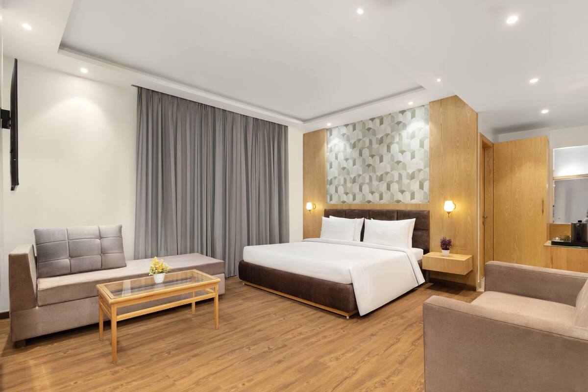 A modern hotel room with a bed, sofa, coffee table, and a small kitchen area.