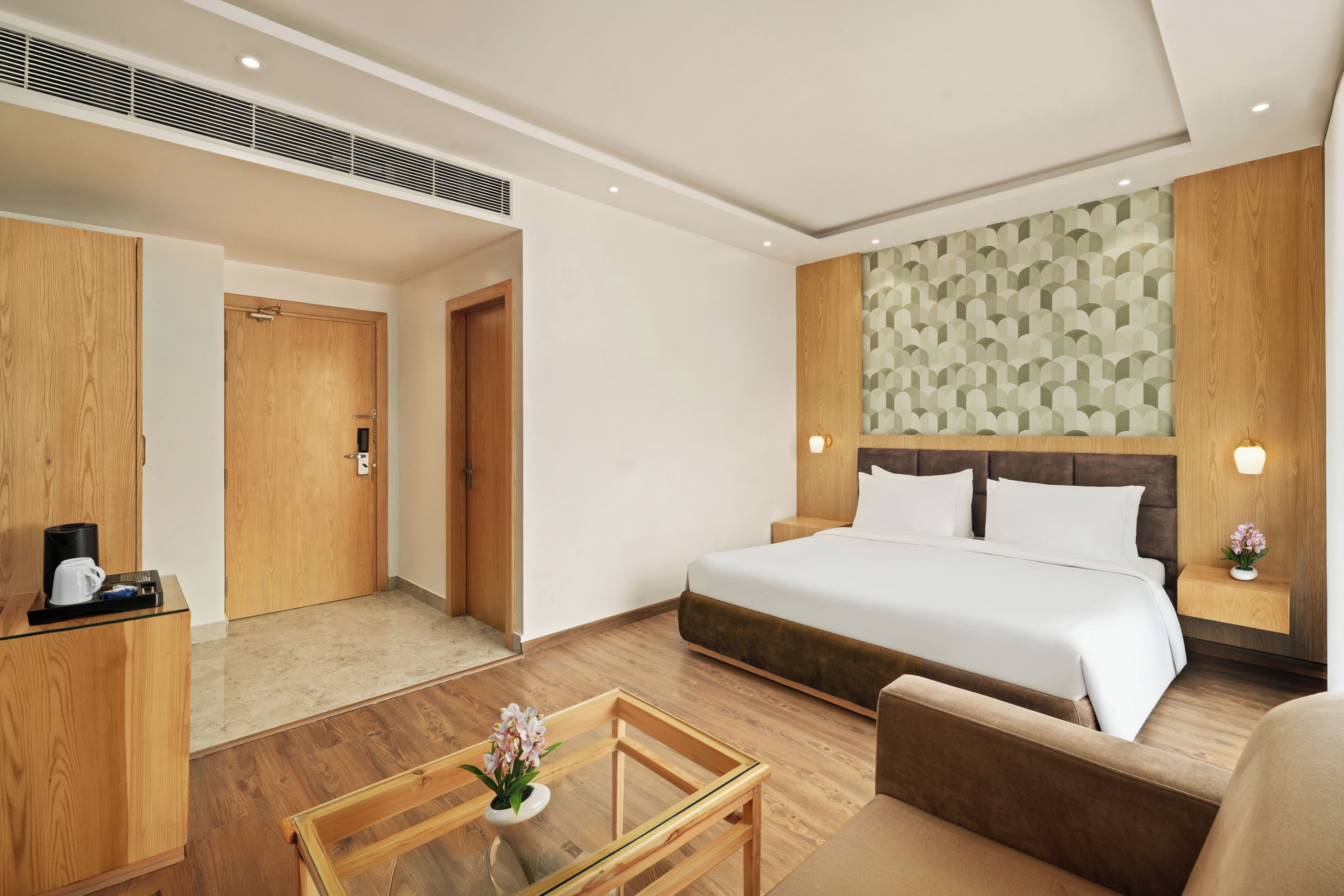 A hotel room with a bed, a desk with a coffee maker, a chair, a small table with a flower, and a patterned wall.