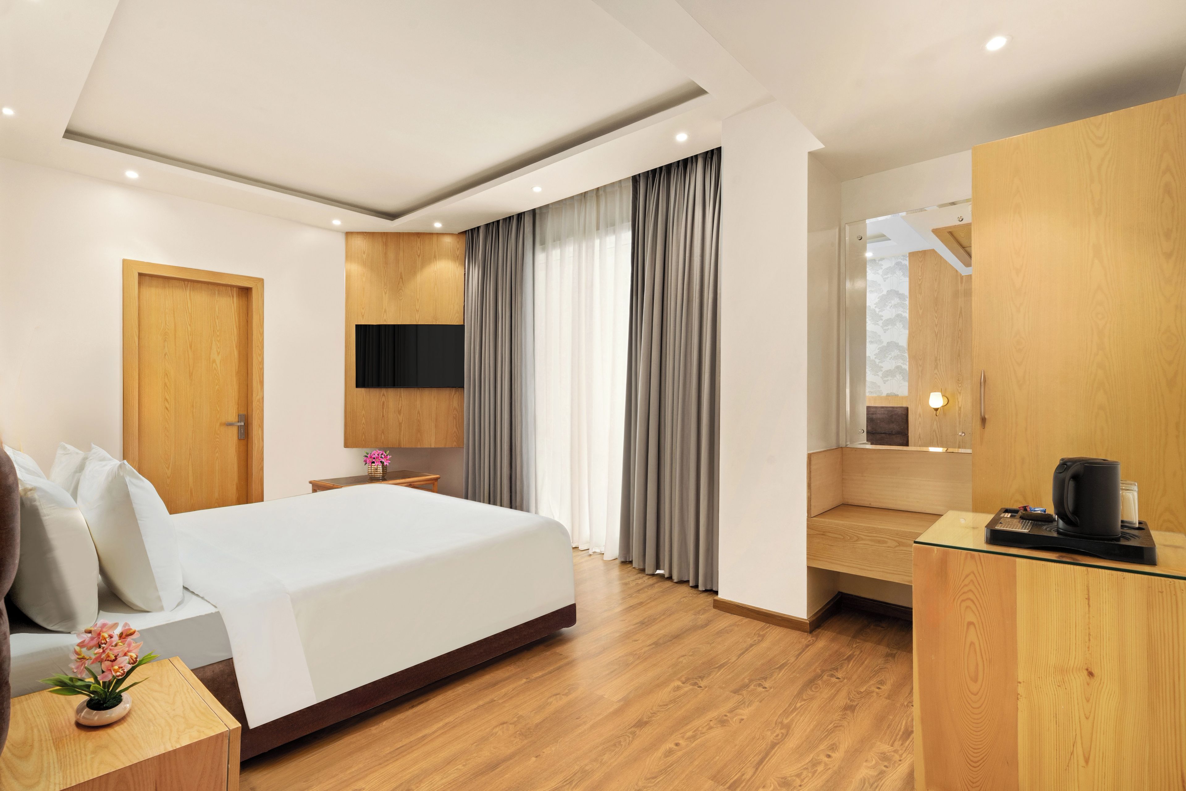 A hotel room with a bed, a desk with a coffee maker, a TV mounted on the wall, and a window with curtains.