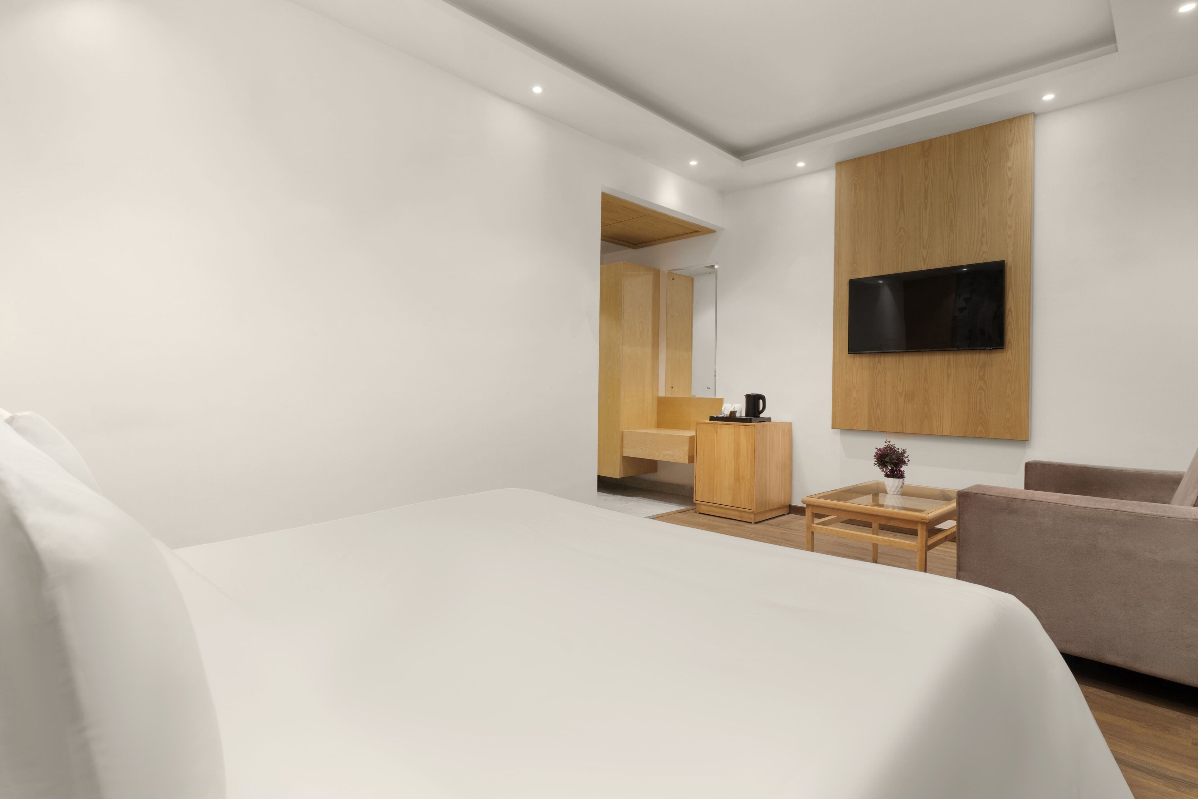 A modern hotel room with a large bed, a sofa, a TV mounted on the wall, a wooden cabinet, and a small table with a plant.