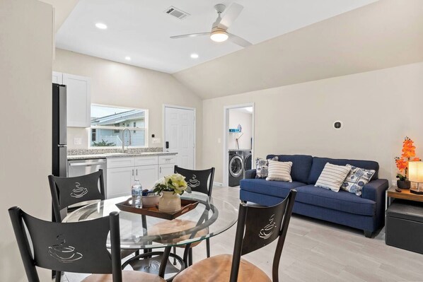 A modern living room with a blue sofa, a dining table with chairs, a ceiling fan, and a kitchen area with a refrigerator and washing machine.