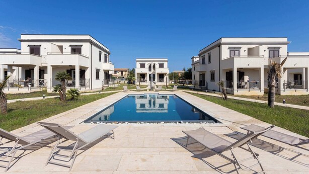 Uma área de piscina com espreguiçadeiras, uma piscina azul cristalina e edifícios modernos com varandas.