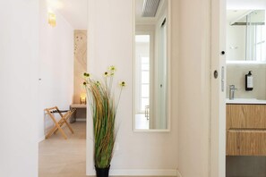 Ein Flur mit einem Blumentopf, einem Wäscheständer aus Holz und einem Spiegel, der einen Raum mit Waschbecken und Holzschränken reflektiert.