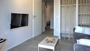 A modern living room with a flat-screen TV, a small wooden coffee table, a grey sofa, and a decorative macrame curtain. - Appartamento "Isola di Panarea 2" (Carini)